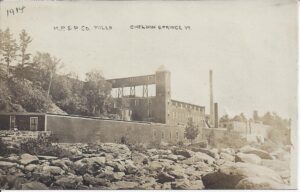 A paper mill along a rocky river bank with two visible chimneys and two long wooden buildings, one is three stories